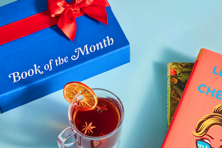 A blue gift box labeled "Book of the Month" next to a cup of tea and colorful books, showcasing cozy reading vibes.