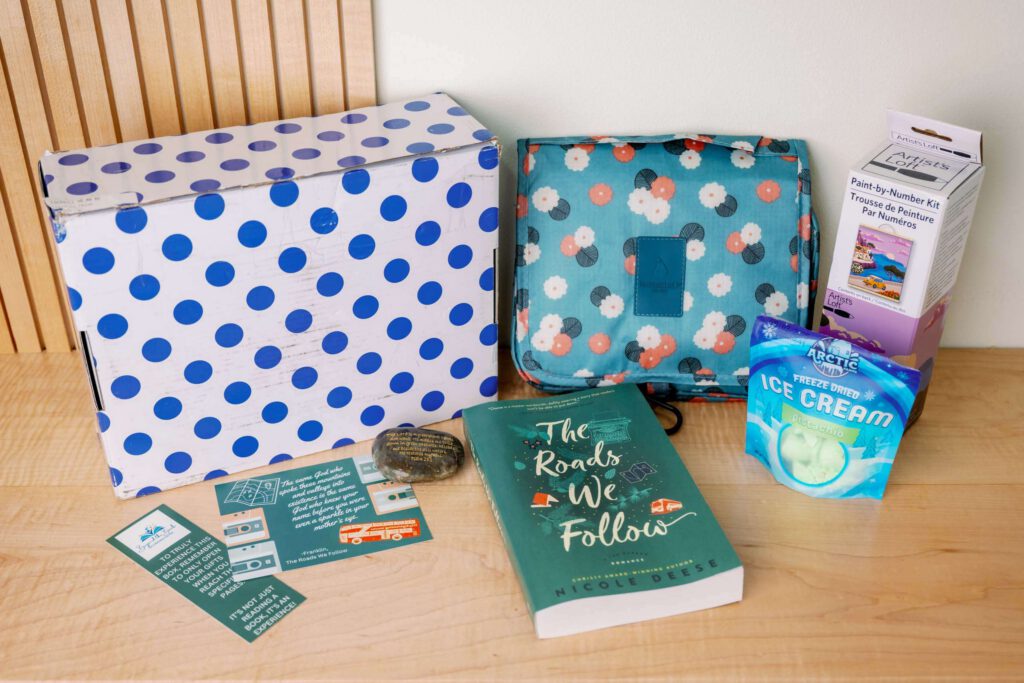 Polka-dot box and contents: book "The Roads We Follow," bookmarks, floral pouch, craft kit, snack — best book subscription boxes