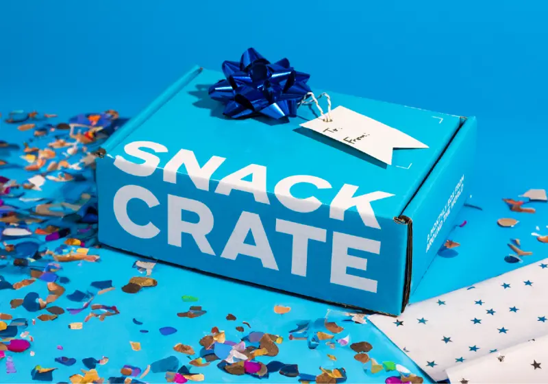 Blue Snack Crate gift box with metallic blue bow and blank tag amid colorful confetti on blue background