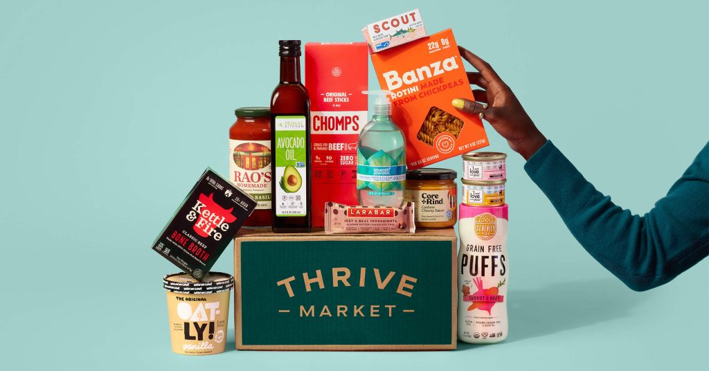 Thrive Market box surrounded by pantry items and snacks, a hand reaching to grab a box of chickpea pasta.