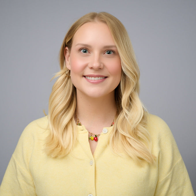 A smiling individual with long, wavy blonde hair wearing a yellow cardigan and a colorful necklace, set against a neutral bac